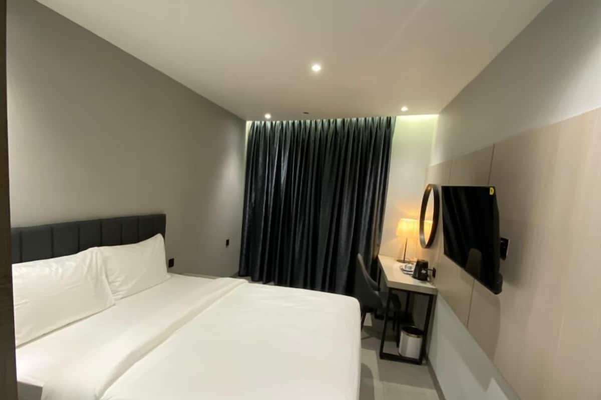 Moderni hotellihuone, jossa on suuri sänky, työpöytä tuolilla, seinälle asennettu taulutelevisio ja ikkuna, jossa on tummat verhot.