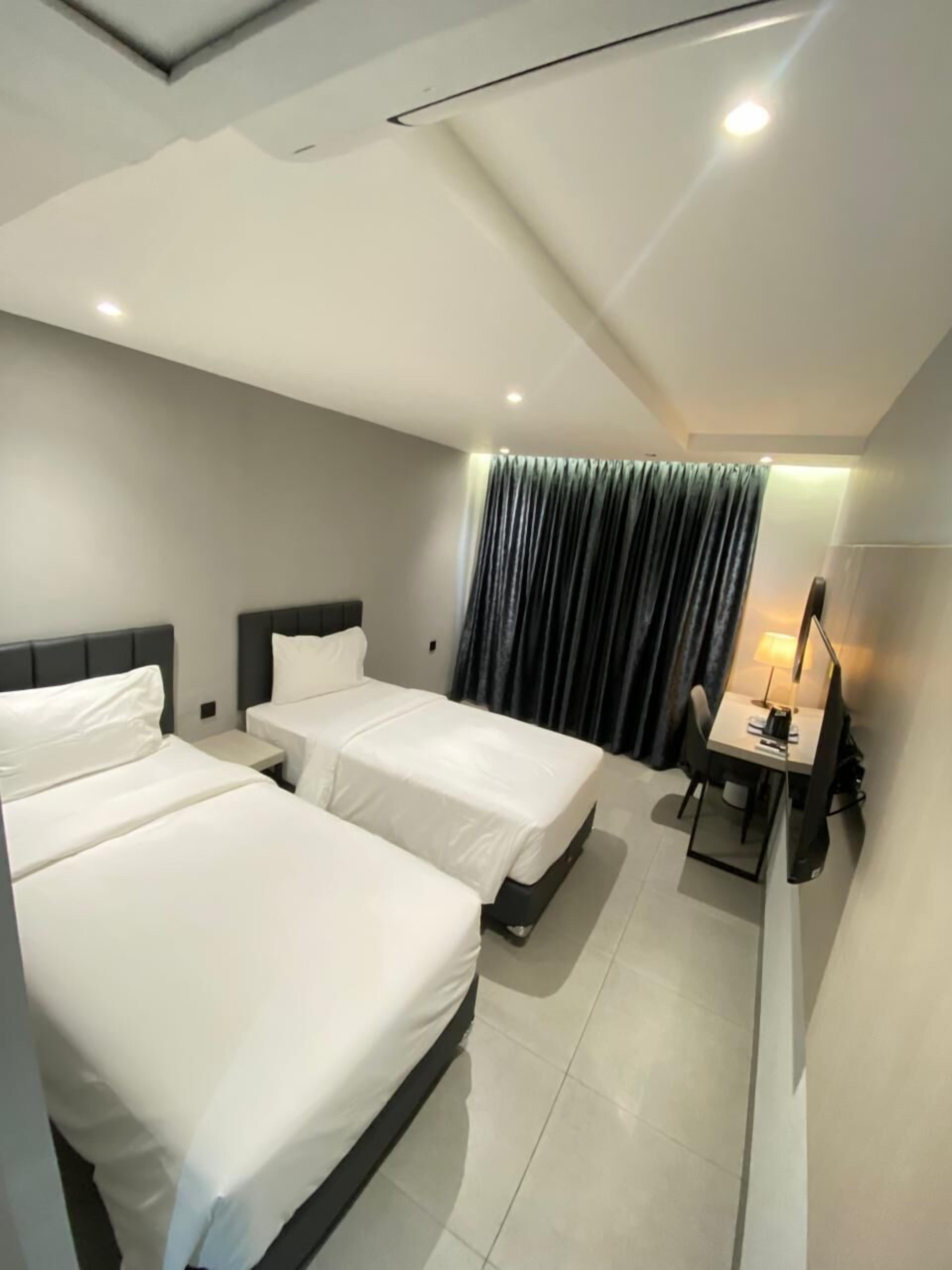 A modern hotel room with two single beds, a desk with a chair, and a large window with curtains.
