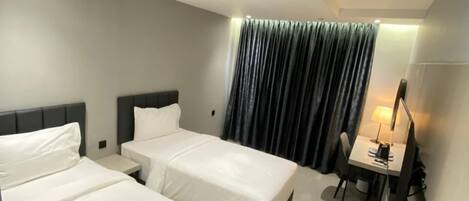 A modern hotel room with two single beds, a desk with a chair, and a large window with curtains.