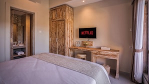 A bedroom with a bed, a wooden wardrobe, a desk with a television displaying 'NETFLIX', and a bathroom visible through an open door.