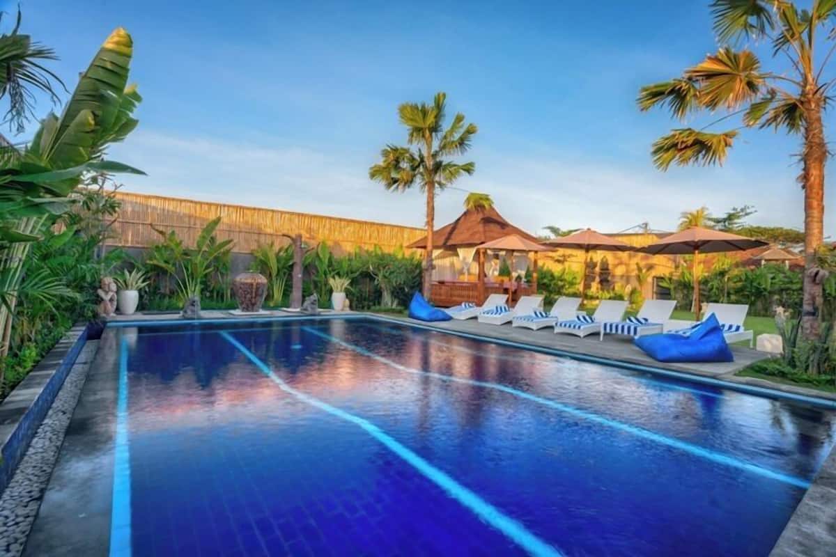A swimming pool with lounge chairs and umbrellas, surrounded by tropical plants and a wooden fence.