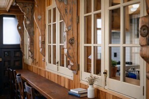 Un interno in legno con un lungo tavolo da pranzo, diverse finestre con stipiti bianchi e un vaso con fiori secchi sul davanzale.