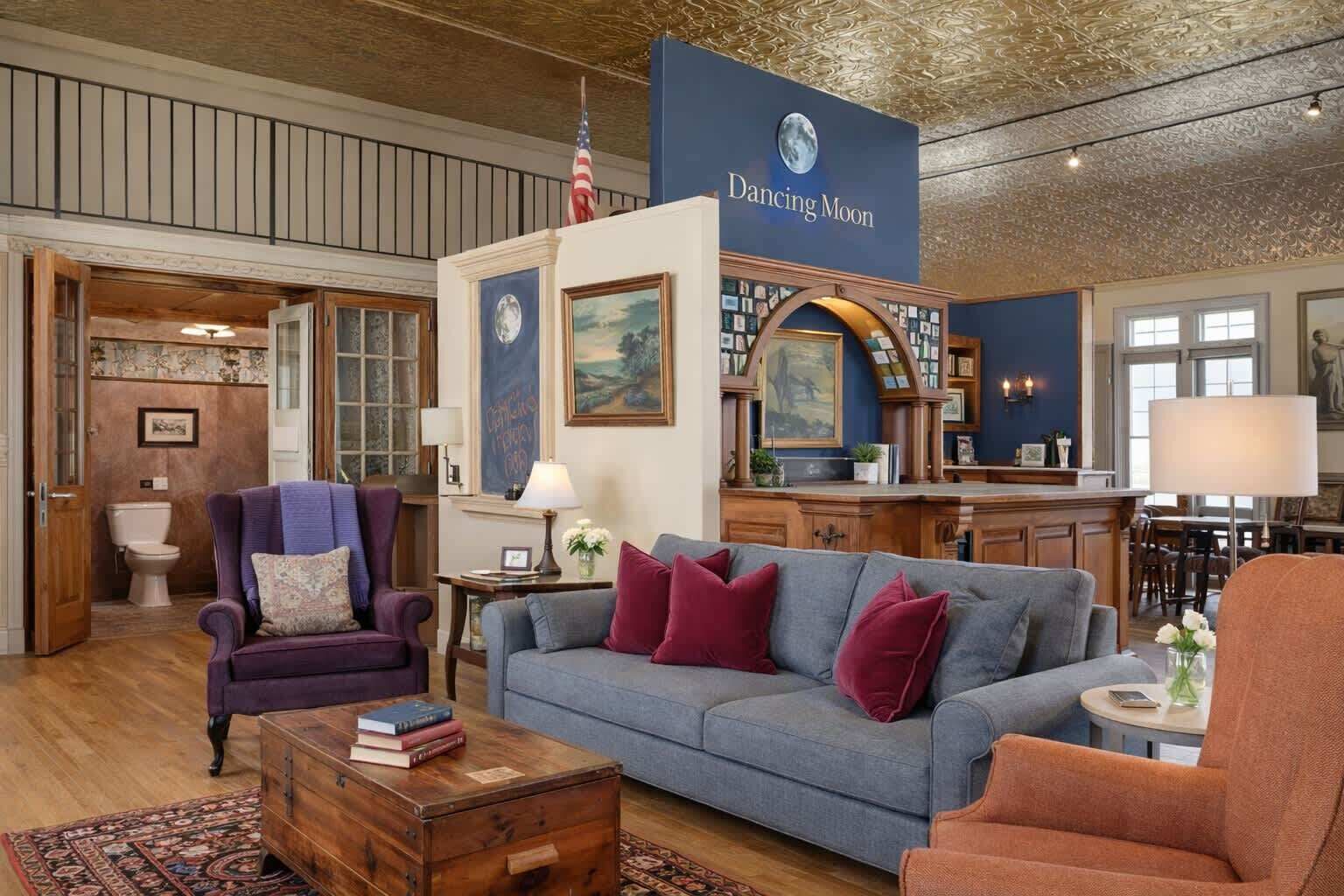 Ein geräumiges Wohnzimmer mit einem blauen Sofa, einem violetten Sessel und einem orangen Sessel. Ein Couchtisch aus Holz mit Büchern befindet sich in der Mitte. Eine große amerikanische Flagge hängt über dem Kamin.