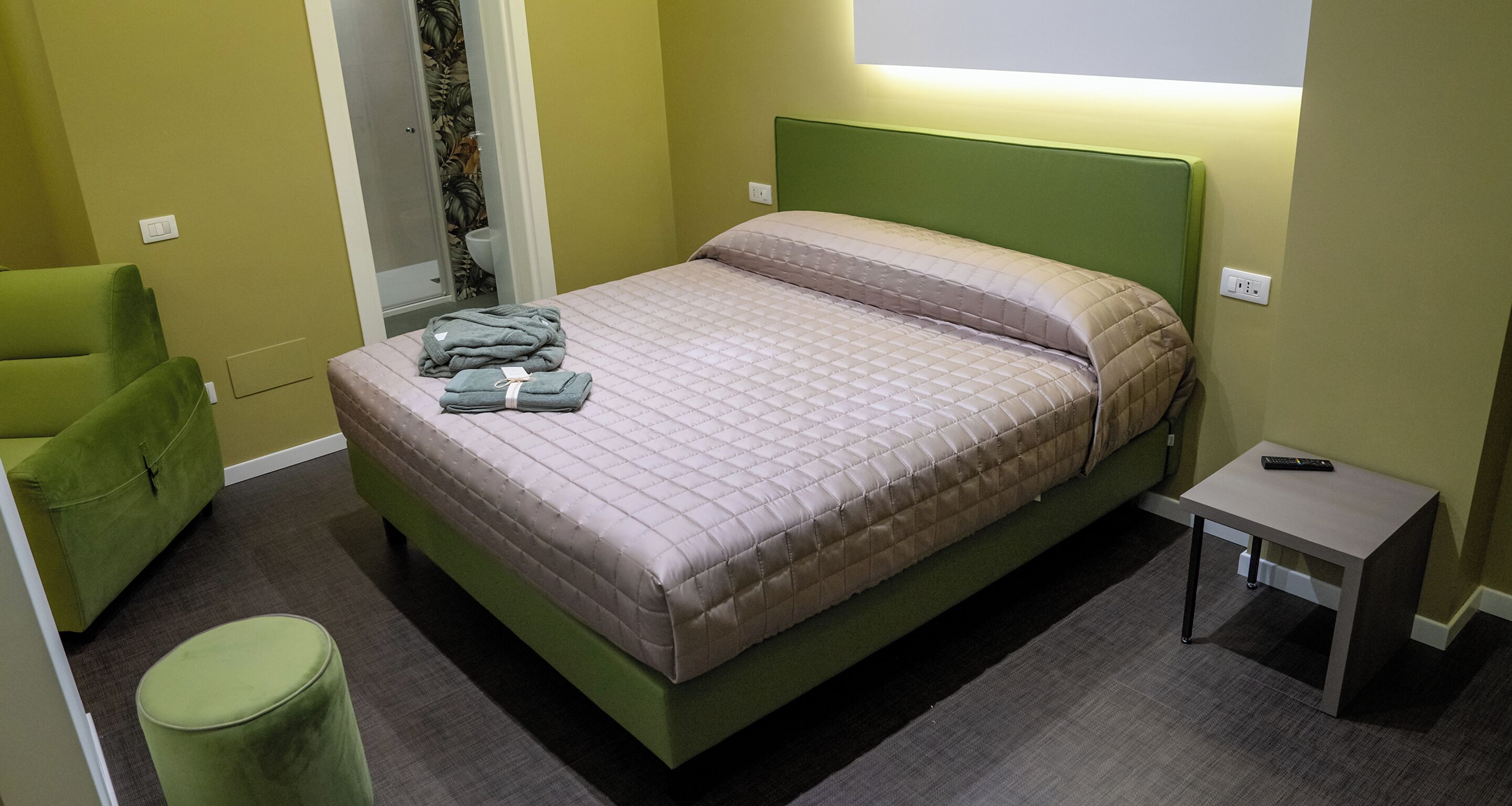 Una cama bien hecha con un cabecero verde, una mesita de noche con un control remoto y una silla verde.
