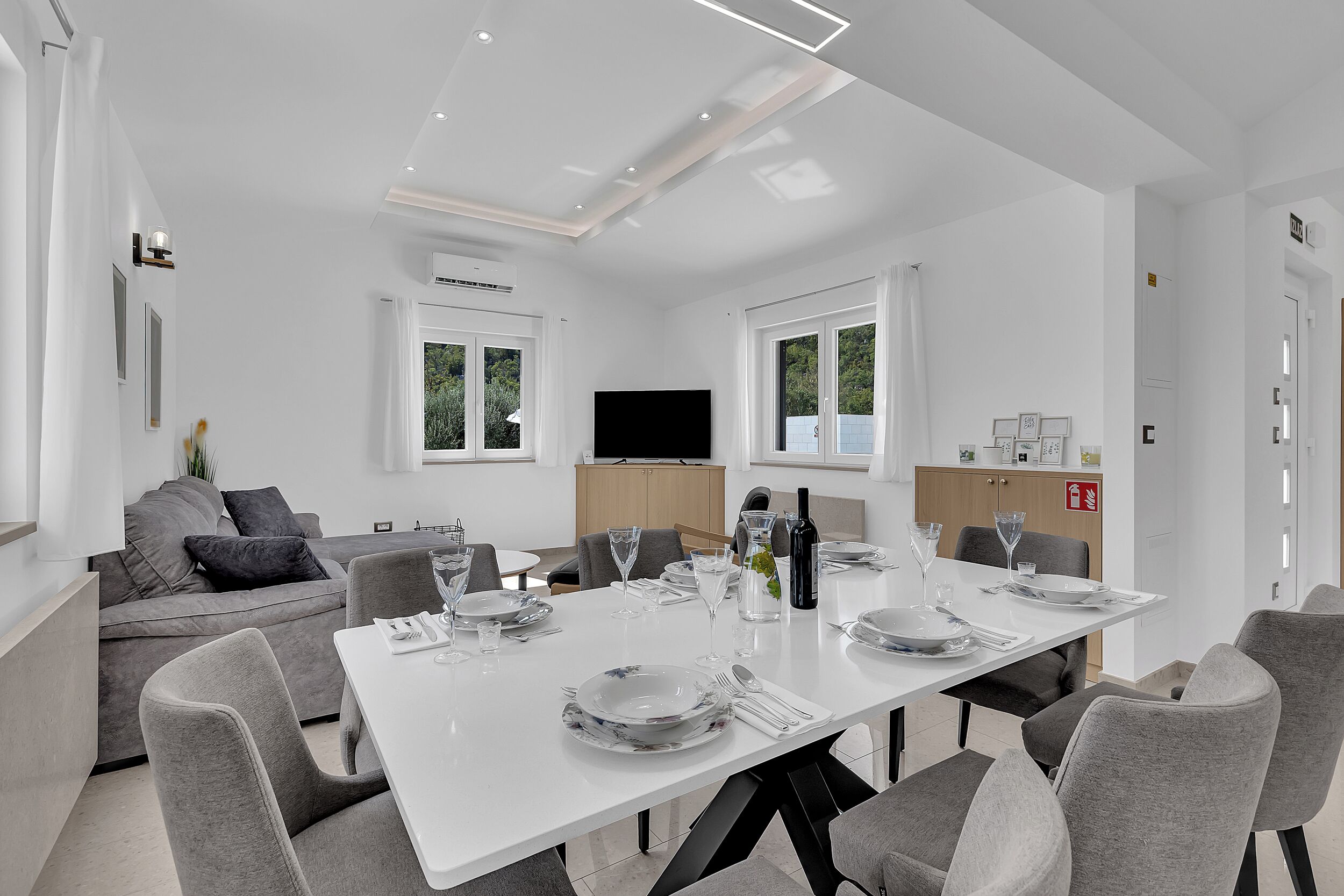 Ein moderner Essbereich mit einem weißen Tisch, gedeckt für sechs Personen, grauen Stühlen und einer grauen Couch im Hintergrund.