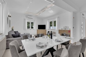 A modern dining area with a white table set for six, grey chairs, and a grey sofa in the background. - Resort Moja Stina - Villa Maslina (Seoca)