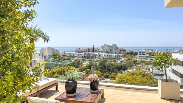 Un balcon avec une table en bois, deux plantes en pot et un canapé. On aperçoit un paysage urbain et la mer en arrière-plan.