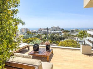 A balcony with a wooden table, two potted plants, and a sofa. A cityscape and the sea are visible in the background.