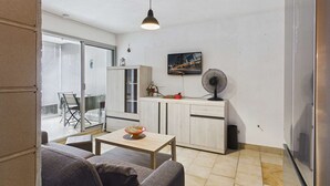 A compact living space with a sofa, a small table, a kitchen area with cabinets, a fan, and a TV mounted on the wall. - 2-room apartment with loggia in Marseillan-Plage (Marseillan-Plage)