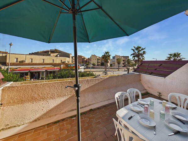 Une terrasse extérieure avec une table dressée pour le repas, un parasol vert et une vue sur des immeubles et des palmiers.