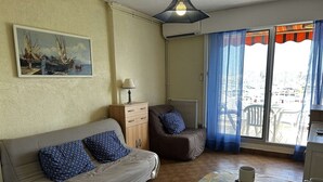 Un salon chaleureux avec un canapé, un fauteuil, une table recouverte d’une nappe à motifs floraux et une vue sur la mer par la fenêtre.