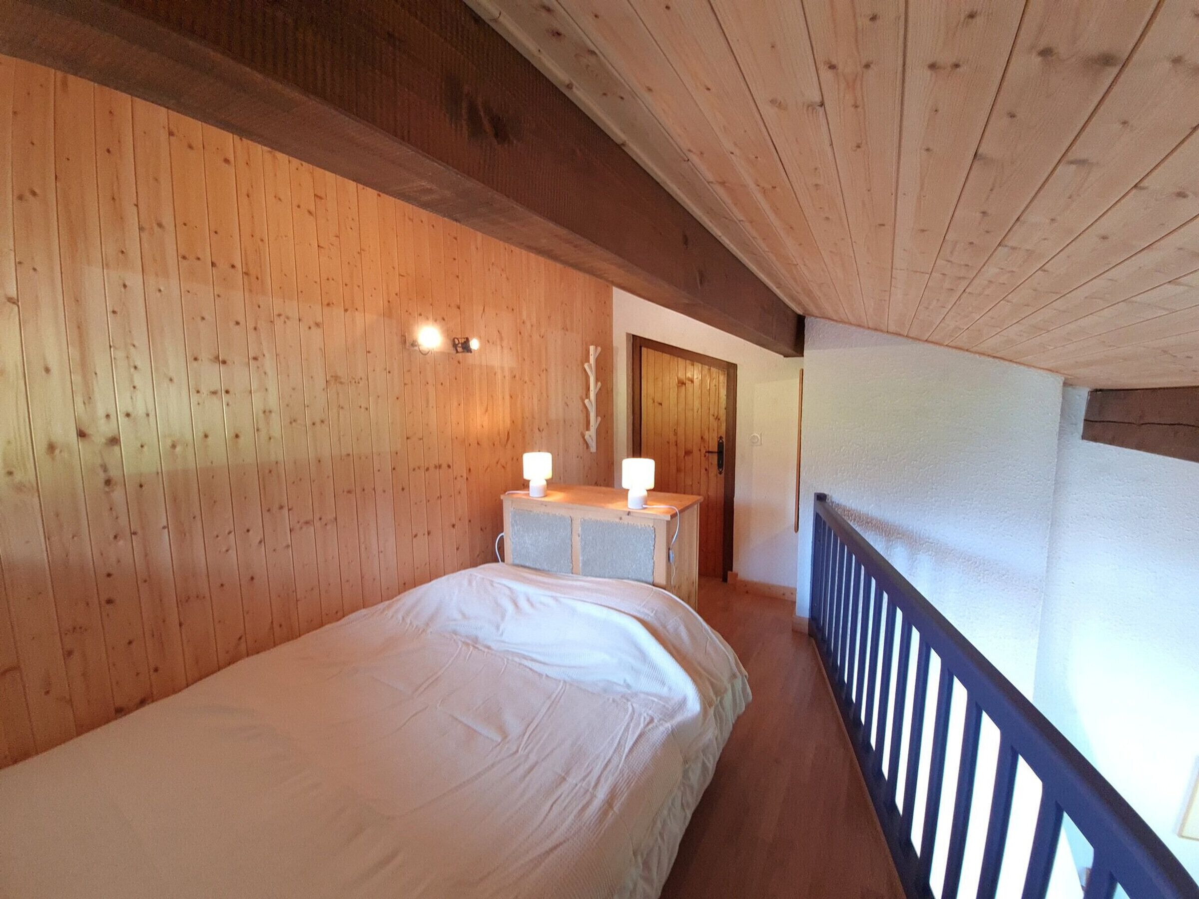 Ein Zimmer mit Holzvertäfelung, einem Bett, einem kleinen Tisch mit Lampe und einer Tür zu einem weiteren Raum.