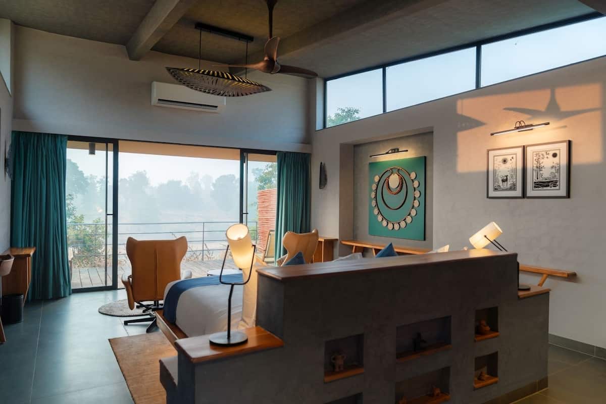 A modern hotel room with a large sliding glass door leading to a balcony, a desk with a lamp, and a wall-mounted art piece.