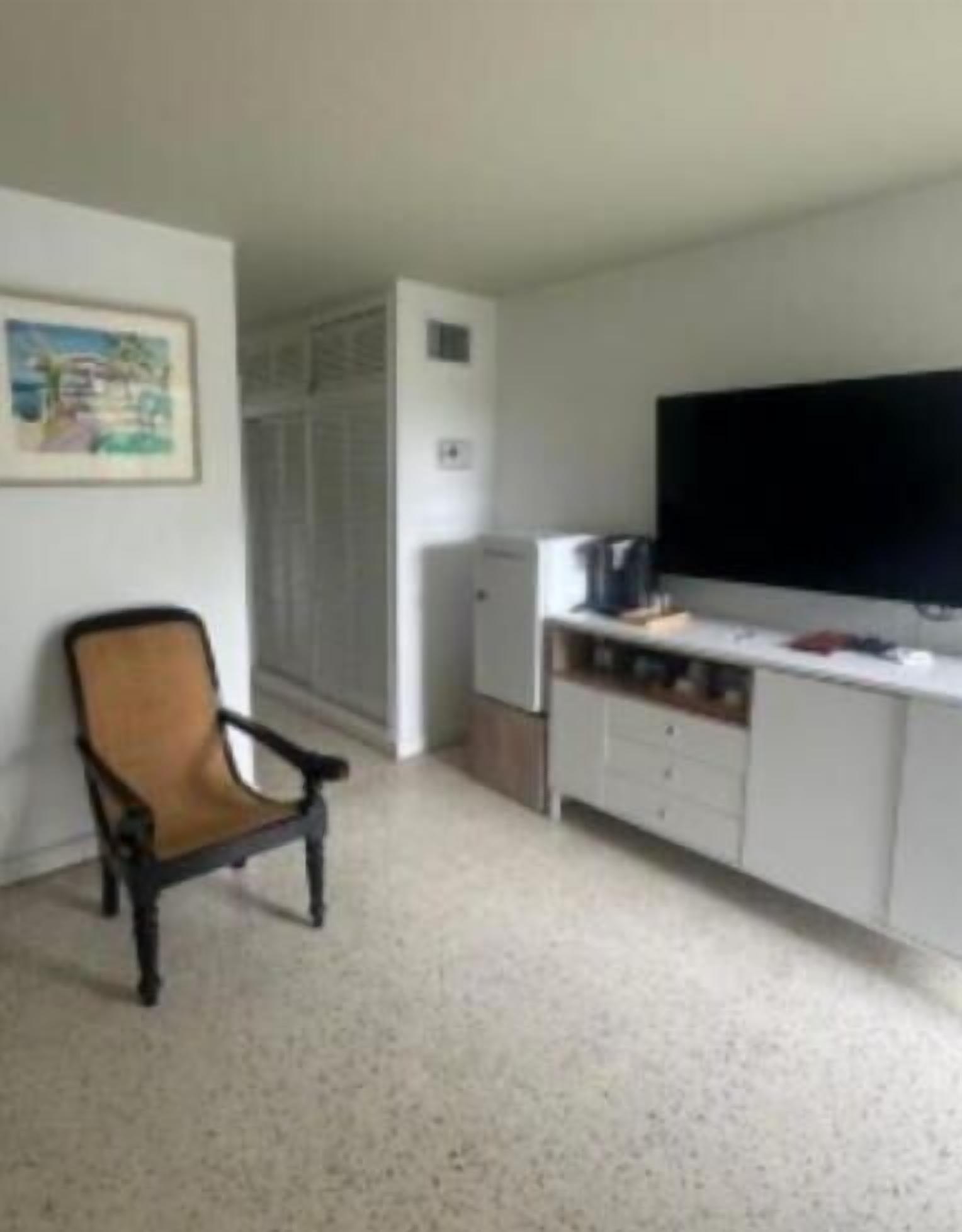 A small, sparsely furnished room with a kitchenette, a single chair, and a large flat-screen TV.