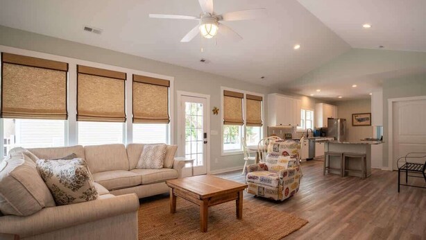 A spacious living room with a sofa, coffee table, and a patterned armchair. There is a kitchen area with a refrigerator and a dining table. - Sea Y'all: Early Bird Special! (Oak Island)