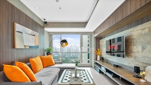 A modern living room with a grey sofa, orange cushions, a coffee table, and a large flat-screen TV mounted on the wall. - Jade by Kozystay - Kuningan (Jakarta)