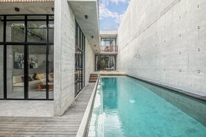 A modern indoor pool area with a clear view of the pool, a wooden deck, and a glass-enclosed seating area with a sofa and cushions.
