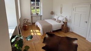 Un dormitorio con un ventanal grande, una cama, una silla, una lámpara y un cuero de vaca en el suelo.