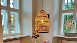 Un comedor con una mesa redonda de madera, sillas rojas, una lámpara colgante de mimbre, un jarrón con flores y una vela.