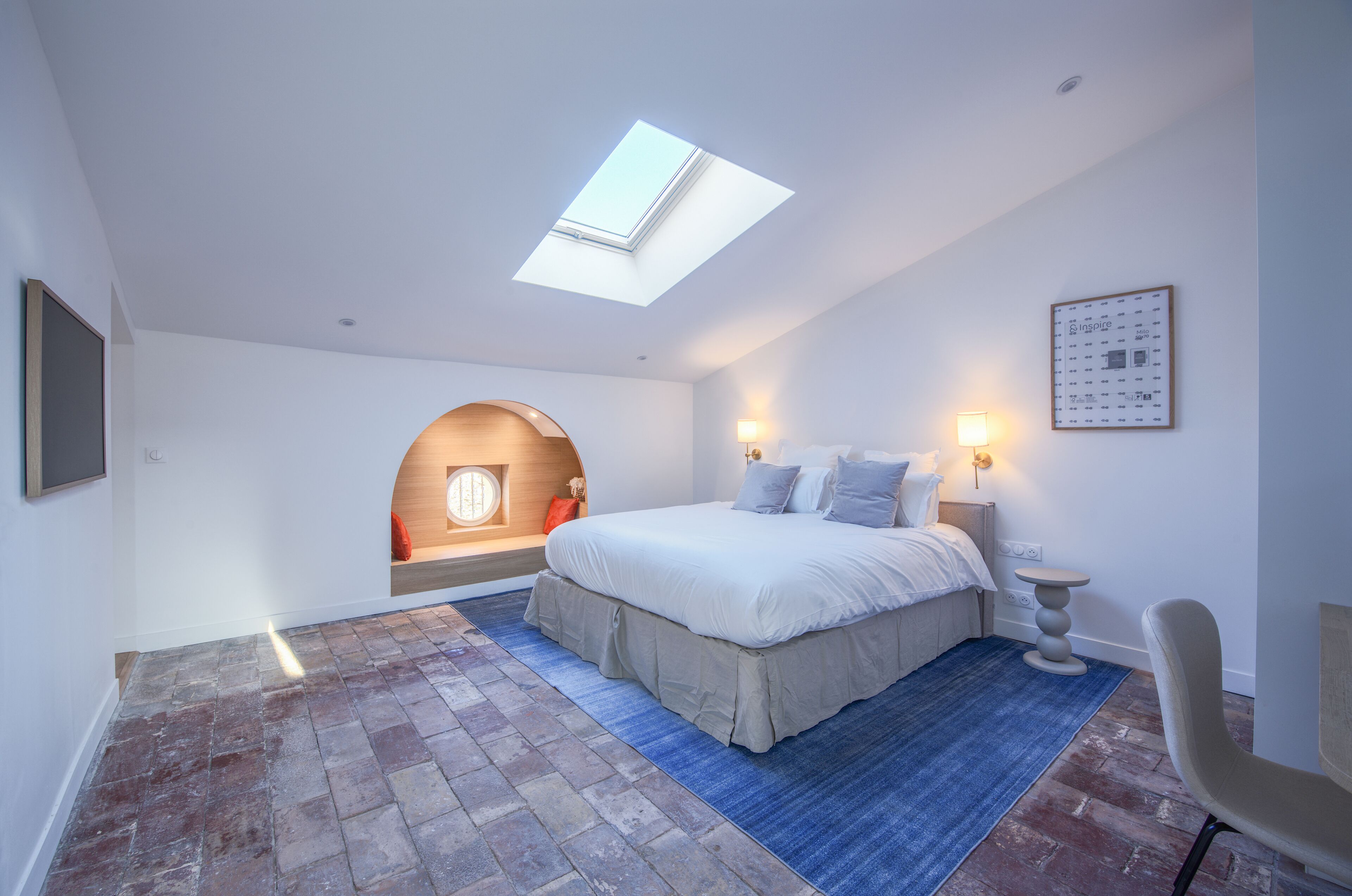 A bedroom with a skylight, a bed with pillows, a nightstand, a chair, and a wall-mounted light fixture.