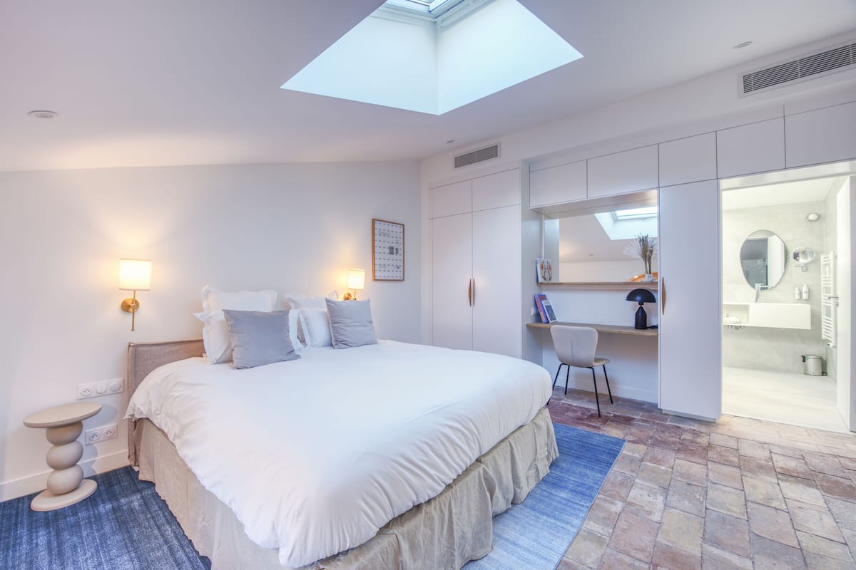 A modern bedroom with a skylight, a large bed, bedside lamps, a desk, and a bathroom with a mirror and sink.