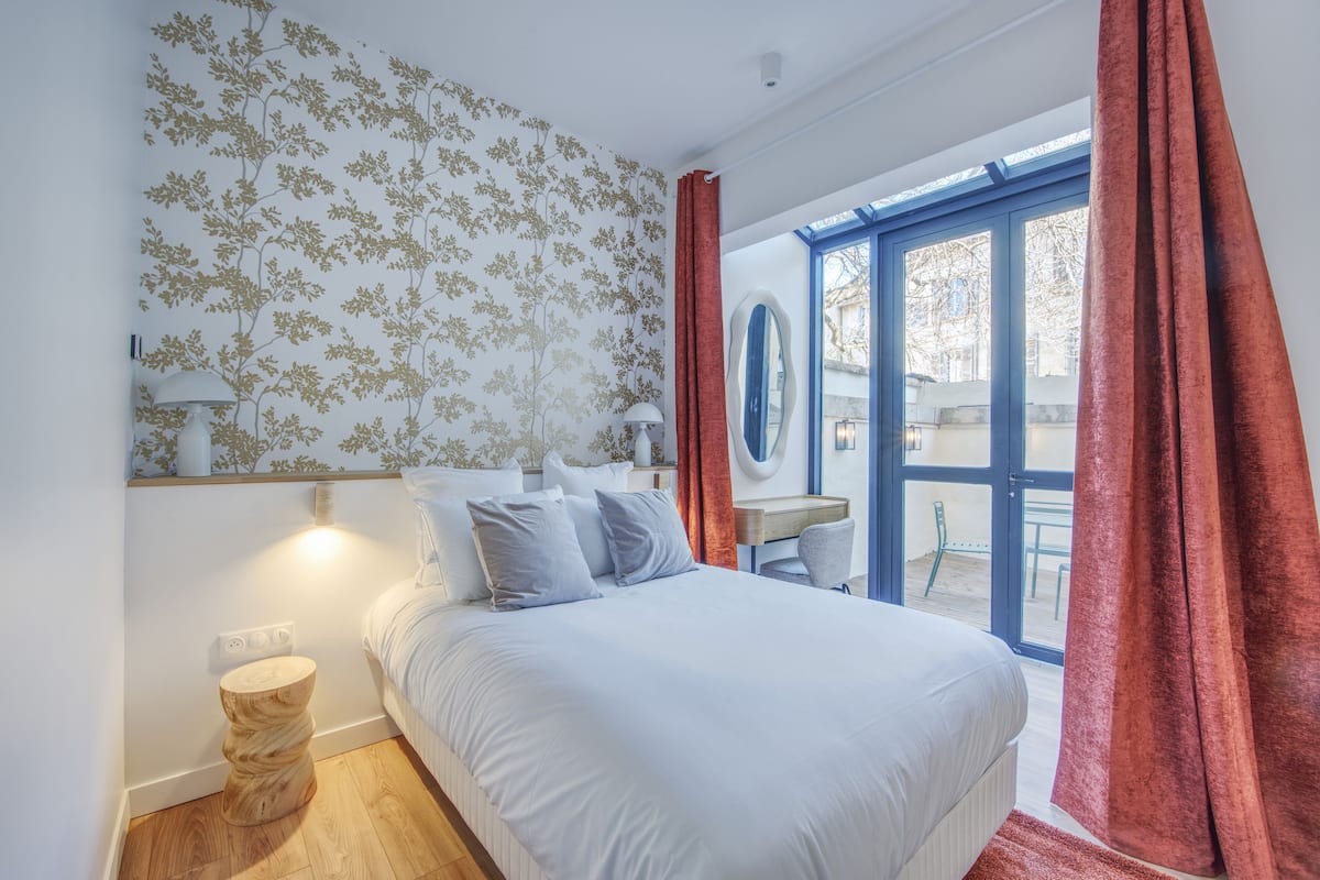 A bedroom with a bed, a nightstand, a mirror, and a window with curtains.