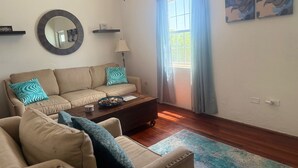 A living room with a beige sofa, a coffee table, and a blue patterned throw. There is a window with blue curtains, a wall-mounted mirror, and two abstract paintings. - Cozy 2-bedroom apartment in enjoyable Sandys with AC, WiFi (Sandys)