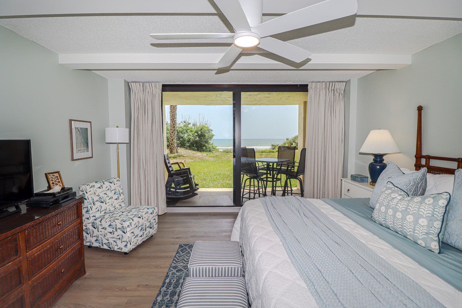 Ein Schlafzimmer mit Bett, Fernseher, einem Holzschrank und Blick auf den Strand durch die Schiebetür.