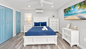 A bedroom with a bed, a ceiling fan, a nightstand, a dresser, a wall-mounted air conditioner, and a beach-themed painting. - Villa Azul | Pool + Guest House | Northwood Hills (West Palm Beach)