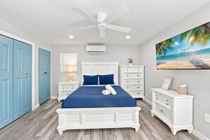 Un dormitorio con cama, ventilador de techo, mesita de noche, cómoda, aire acondicionado de pared y un cuadro con motivos playeros.