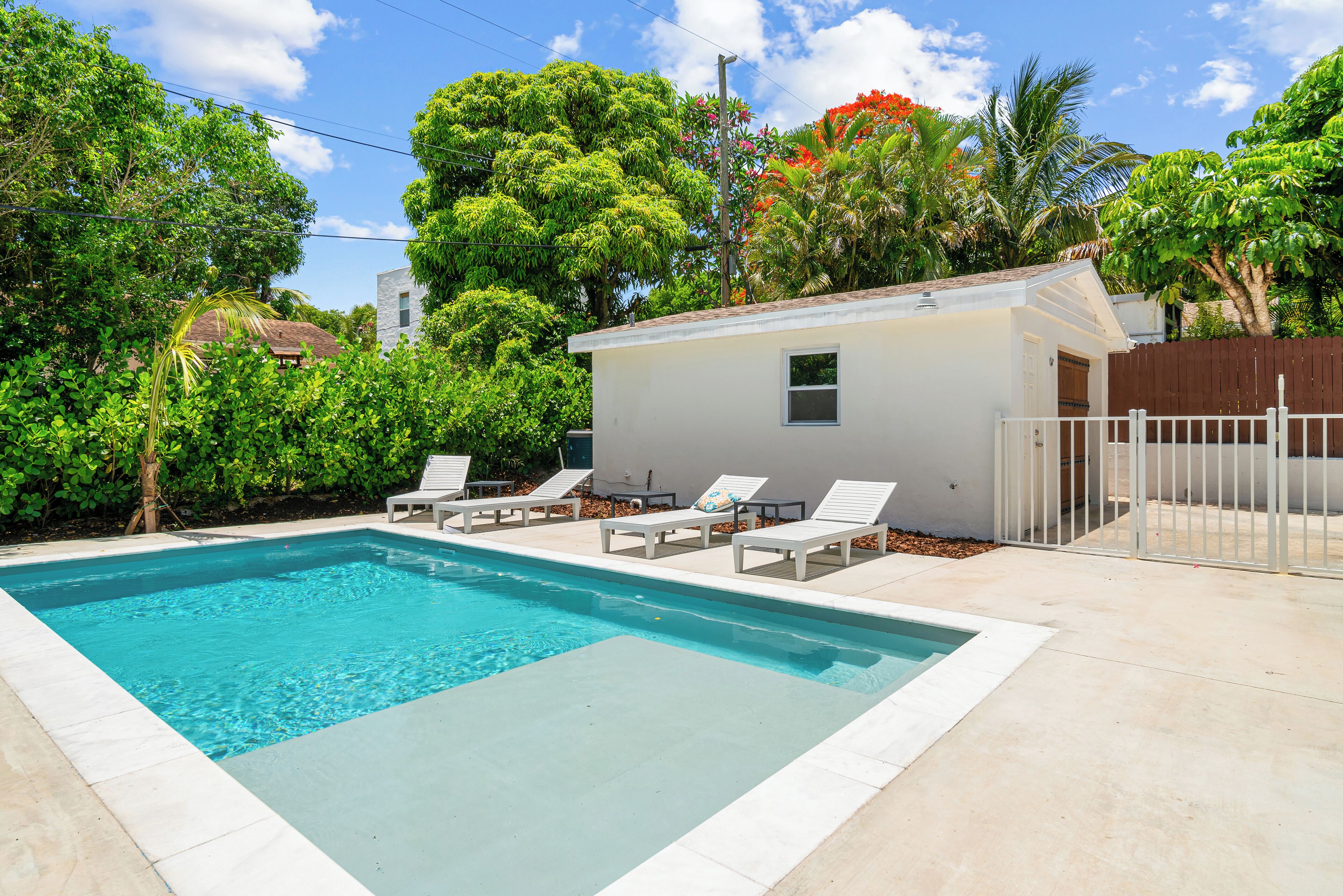 Uma piscina cercada por vegetação exuberante e um pequeno edifício branco, com cadeiras de espreguiçadeira brancas.