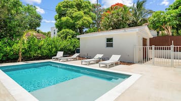 Uma piscina cercada por vegetação exuberante e um pequeno edifício branco, com cadeiras de espreguiçadeira brancas.