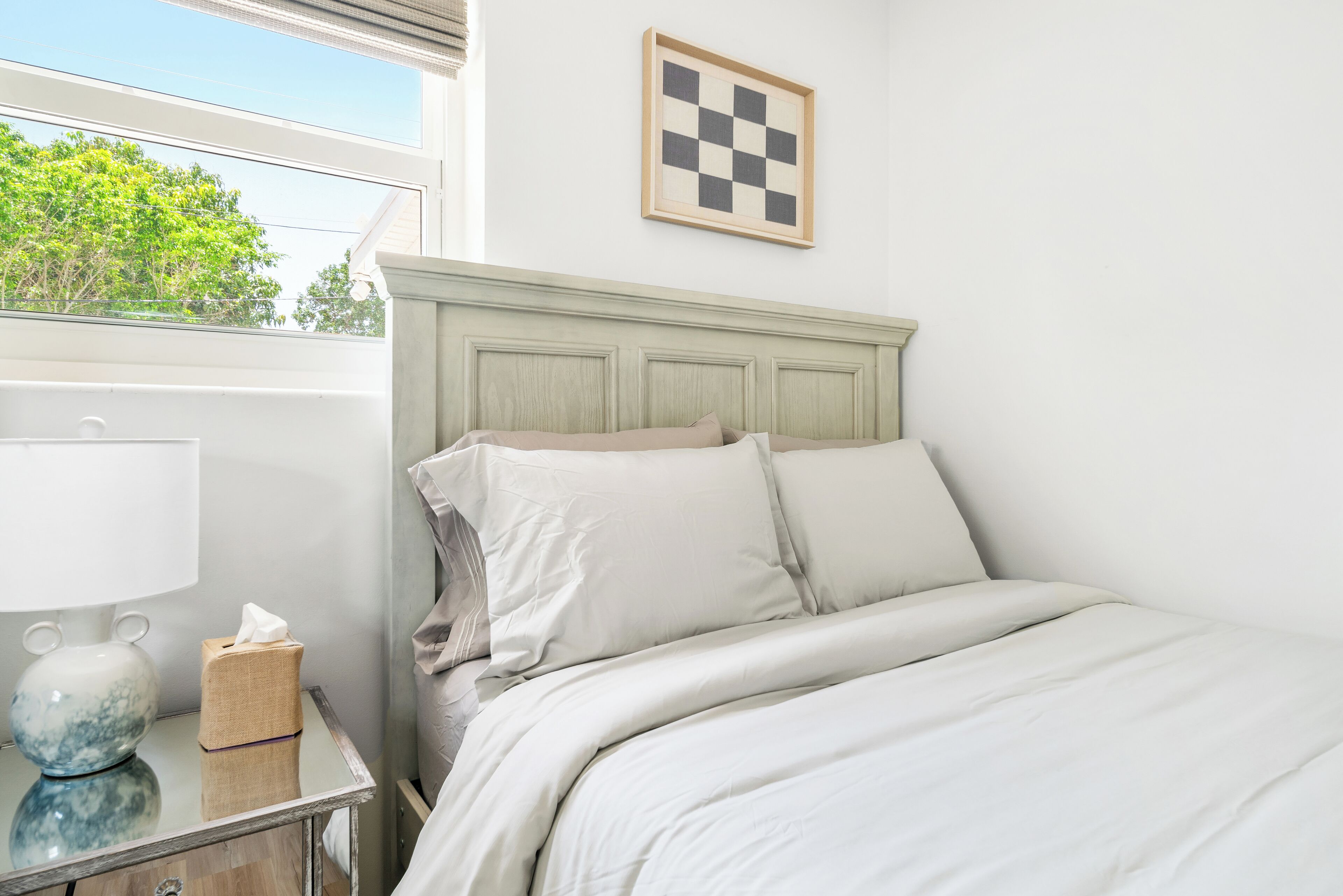 Cama bem arrumada com roupa de cama branca, luminária de cabeceira, caixa de lenços e um quadro de parede xadrez.