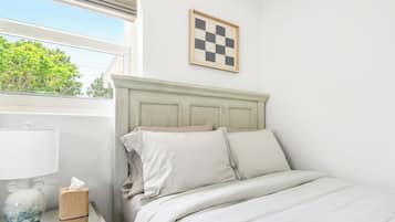 Cama bem arrumada com roupa de cama branca, luminária de cabeceira, caixa de lenços e um quadro de parede xadrez.