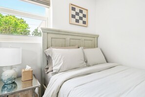 Una cama bien hecha con ropa de cama blanca, una lámpara de noche, una caja de pañuelos y un cuadro de pared a cuadros.