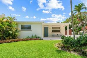 Una casa de dos pisos con un porche delantero, un pequeño jardín y un cielo azul despejado.