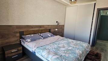 A modern bedroom with a wooden headboard, a bed with floral bedding, a bedside table, and a hanging light fixture.