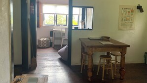 A narrow hallway leading to a dining area with a wooden table and two stools, a window with blinds, and a wall-mounted poster.