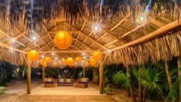 Una estructura interior de madera con techo de paja, múltiples luces esféricas colgantes y un área de asientos central.