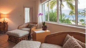 Una habitación con cama, dos sillas, una mesita, una lámpara y una ventana que da a palmeras y al océano.
