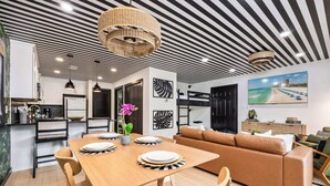 A modern living room with a wooden dining table, brown leather sofa, and a large painting of a beach. - 30A Condo · Walk to Beach · 2 Pools + Tennis (Santa Rosa Beach)