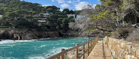 Un camino costero con barandilla de piedra que lleva a una costa rocosa con aguas turquesas.