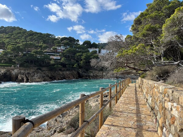 A coastal walkway with a stone railing, leading to a rocky shoreline with turquoise waters.