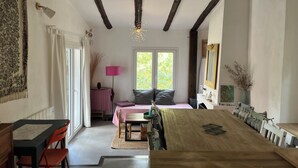 A cozy room with a wooden dining table, a sofa with cushions, a small side table with a lamp, and a window with a view of greenery.
