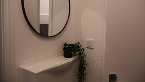 A bathroom with a white wall, a round mirror, a white shelf, and a potted plant.