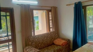 A room with a patterned sofa, a bed with a floral bedspread, a window with a blue curtain, and an air conditioning unit. - Mount Valley Mama Cottage (Rishikesh)