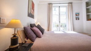 Un dormitorio con cama, mesita de noche, lámpara, ventana con cortinas y un cuadro en la pared.