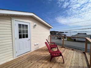 A wooden deck with two red chairs, a white door, and a view of a body of water and buildings in the background. - Iceberg Lookout (Twillingate)