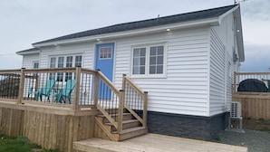 A small, single-story house with a blue door, a wooden deck, and a staircase leading to the front door. - Spiller's Cottage (Twillingate)