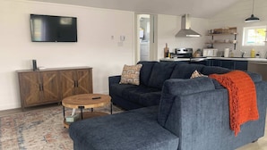 A living room with a sectional sofa, a round coffee table, a TV mounted on the wall, a wooden cabinet, and a kitchen area visible through an open doorway. - Spiller's Cottage (Twillingate)